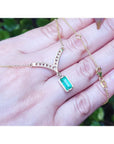 14K yellow gold necklace with a baguette-cut Colombian emerald dangling from a diamond-studded curved V, shown resting on a person's hand against a blurred green background.