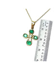 Yellow gold cross pendant with four round emeralds and a diamond center, shown with a ruler.