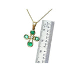 Yellow gold cross pendant with four round emeralds and a diamond center, shown with a ruler.