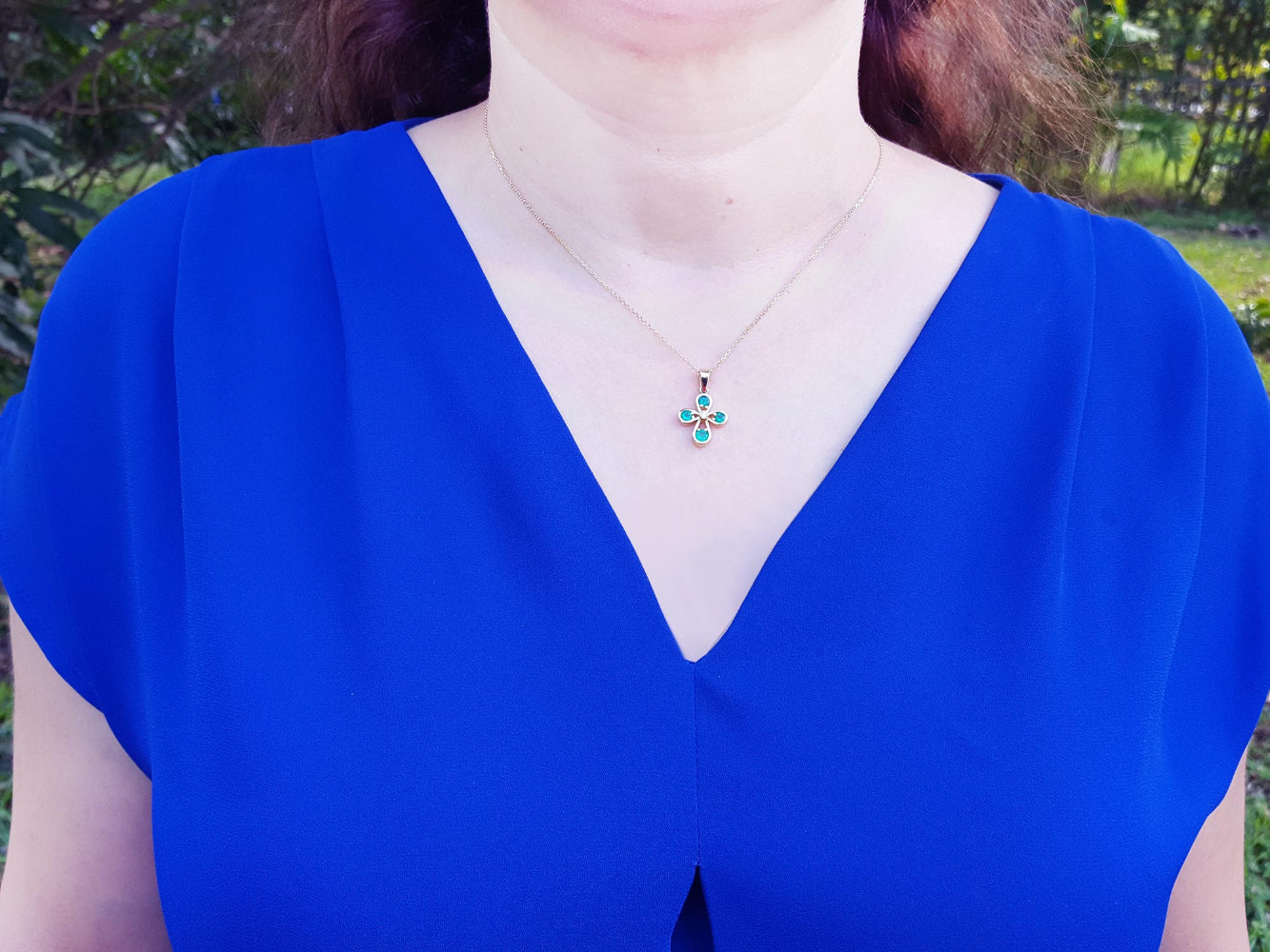 A woman wearing a blue top with a necklace featuring a cross emerald pendant outdoors.