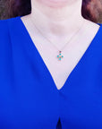 A woman wearing a blue top with a necklace featuring a cross emerald pendant outdoors.