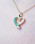 Heart-shaped pendant with emeralds and a diamond on a gold chain against a pink background