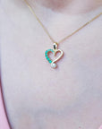 Heart-shaped pendant with natural emeralds on a gold chain, worn by a person against a pink background.