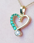 Heart-shaped gold pendant with emerald gemstones on a light pink background.