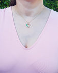 A woman wearing a light pink blouse and a heart emerald pendant with a blurred green background.