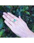 Hand wearing a white gold ring for men with a natural colombian emerald against a leafy background.
