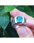 A bezel set natural colombian emerald solitaire ring for men held by a hand against a green leafy background.