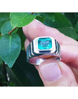 Men's 18K white gold ring with a deep bluish-green, bezel-set, modified emerald-cut Colombian emerald.