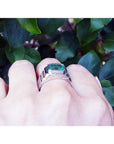 A men's emerald solitaire ring white gold is on a hand against a leafy background.