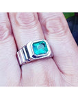 A 18k white gold men's ring with a bezel set natural colombian emerald worn on a finger.