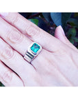 A men's white gold solitaire emerald ring on a hand against a blurred natural background.