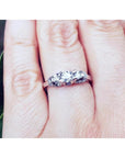14K white gold three-stone round cut diamond engagement ring with pave accent diamonds on a hand.