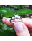 14K white gold three-stone princess cut diamond ring held by fingers with green leaves in background.
