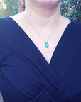 A woman wearing a blue dress with a necklace featuring a natural emerald pendant outdoors.