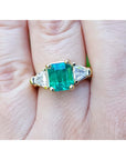 A Gia certified emerald ring in 18k yellow gold and trillion cut side diamond accents on a finger.