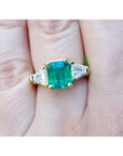 A Gia certified emerald ring in 18k yellow gold and trillion cut side diamond accents on a finger.