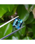 GIA certified 6.07 ct. rectangular emerald-cut blue-green sapphire held by tweezers against green leaves.