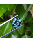 GIA certified 6.07 ct. rectangular emerald-cut blue-green sapphire held by tweezers against green leaves.
