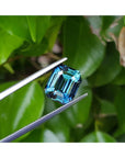 GIA certified 6.07 ct. rectangular emerald-cut blue-green sapphire held by tweezers against green leaves.