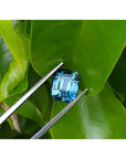 GIA certified 6.07 ct. rectangular emerald-cut blue-green sapphire held by tweezers on green leaves.