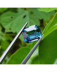 GIA certified 6.07 ct. rectangular emerald-cut blue-green sapphire held by tweezers against green leaves.