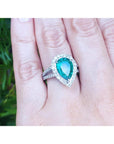 Ring with a teardrop emerald and halo diamond on a hand against a blurred green background.