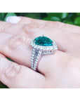Emerald ring with diamonds on a finger against a blurred green background.