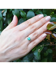 14K yellow gold emerald ring with baguette side diamonds on a hand, against green leaves.