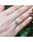 Hand wearing a, emerald May's birthstone ring against a leafy background.