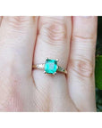 A yellow gold emerald ring with side diamonds on a person's finger, with a natural background.