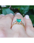 Gold ring with a natural emerald on a hand against a leafy background.