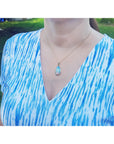 A woman wearing a blue and white patterned top with a gold necklace featuring an opal pendant.