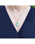 Emerald-cut emerald pendant with halo diamonds on an 18K gold chain, worn on a dark blue top.