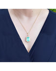 Emerald-cut emerald pendant with halo diamonds on an 18K gold chain, worn on a dark blue top.