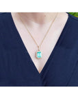 Emerald-cut emerald pendant with halo diamonds on an 18K gold chain, worn on a dark blue top.