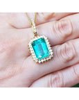Emerald-cut emerald pendant with halo diamonds in 18K gold, worn on a hand.