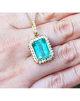 Emerald-cut emerald pendant with halo diamonds in 18K gold, worn on a hand.