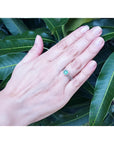 Hand wearing a 14K white gold emerald and halo diamond engagement ring against green leaves.
