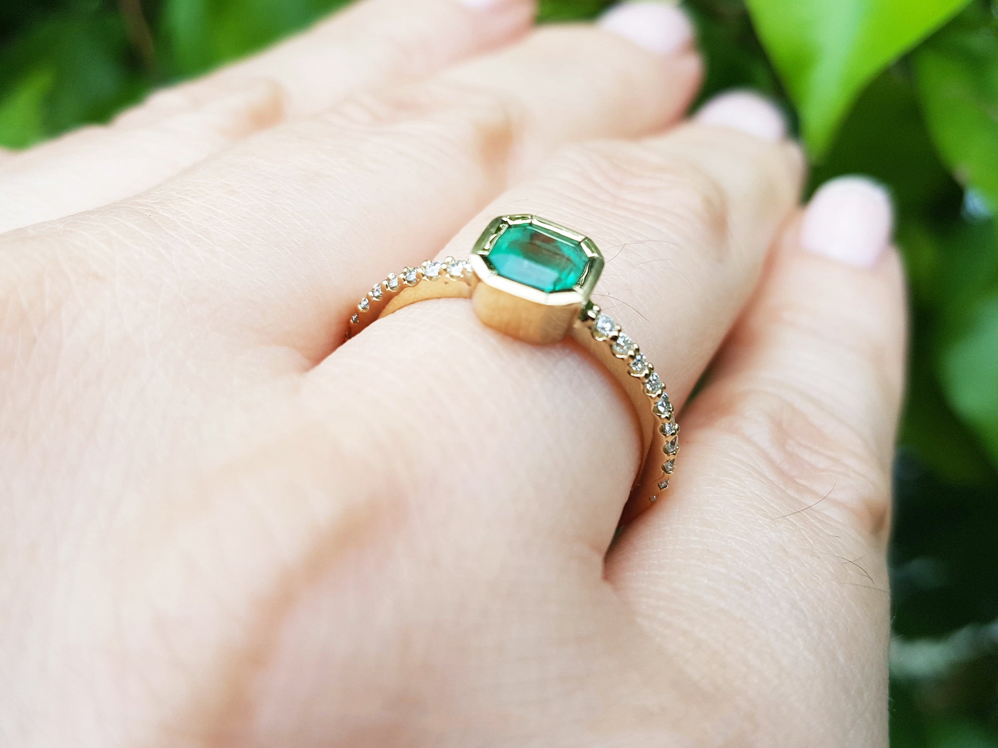 Gold ring with a natural emerald on a hand against a blurred green background.