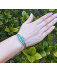 14k white gold bangle bracelet with round Colombian emeralds and diamonds on a hand against green leaves.