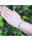 A woman's hand wearing a gold bracelet with five high quality colombian emeralds against a green leafy background.
