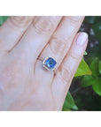 Hand wearing a white gold ring with a blue sapphire against a green leafy background