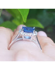 Asscher cut blue sapphire ring with halo diamonds in 18k white gold, worn on a finger.