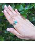 Hand wearing a white gold mens ring with a asscher cut natural colombian emerald against a green leafy background.