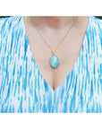 A woman wearing a blue and white patterned top with a gold necklace featuring a natural opal pendant.