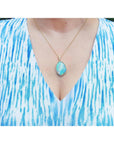 A woman wearing a blue and white patterned top with a gold necklace featuring a natural opal pendant.