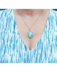 A woman wearing a blue and white patterned top with a gold necklace featuring a natural opal pendant.