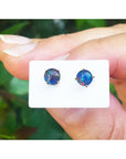 Opal stud earrings on a white card held between fingers with a blurred green background.