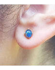 Round Australian opal doublet stud earring with blue and green colors, prong-set in 14K yellow gold on an earlobe.