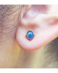 Round Australian opal doublet stud earring with blue and green colors, prong-set in 14K yellow gold on an earlobe.